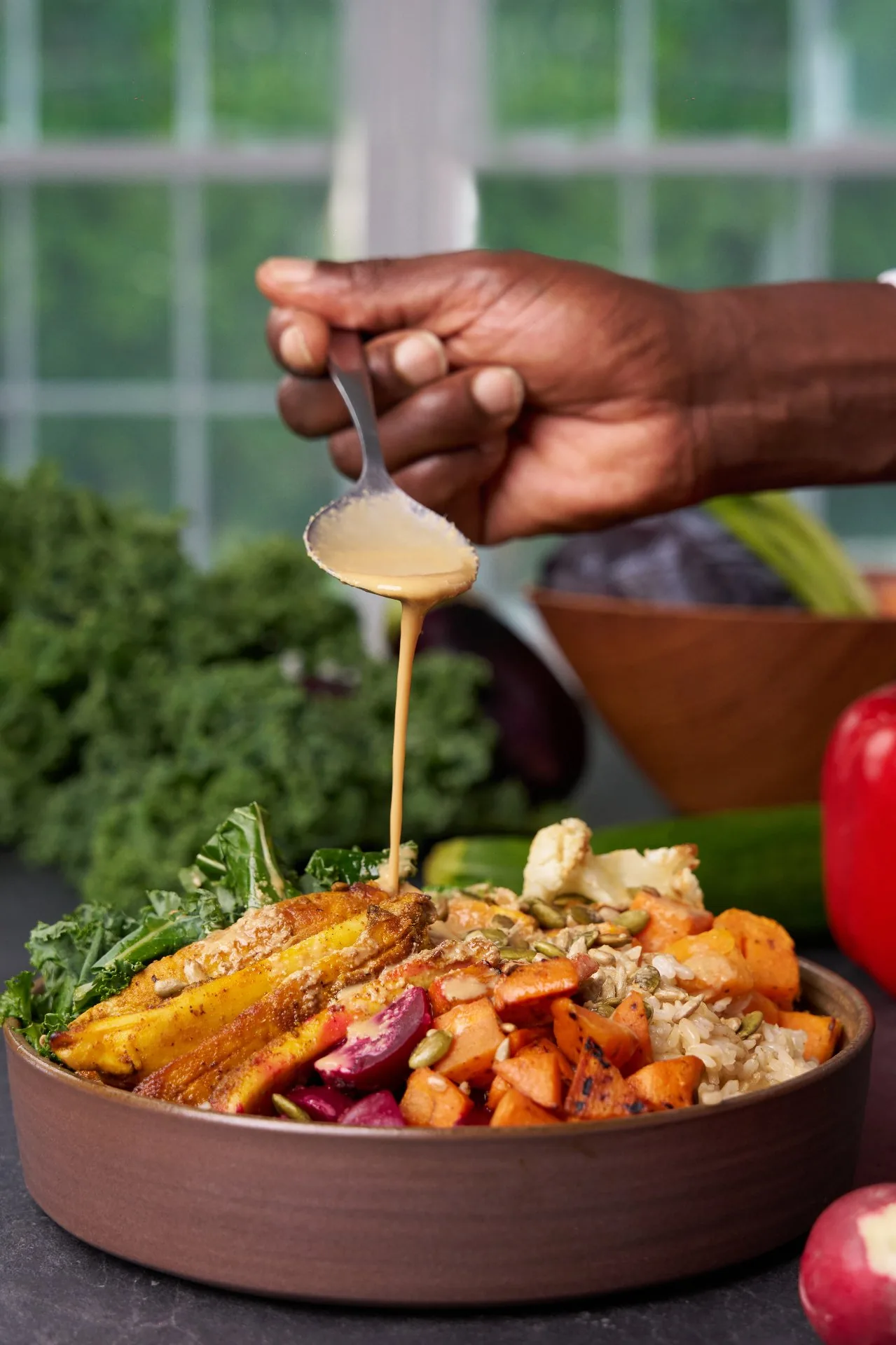 Hand drizzling sauce over a healthy bowl with grilled plantain, roasted vegetables, brown rice, kale, and fresh produce